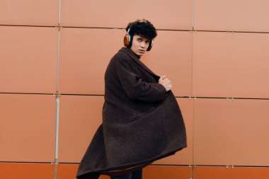 Paltolu ve kulaklıklı genç bir adamın moda fotoğrafı turuncu bir duvarın önünde duruyor, arka planda kırmızı bir duvar var..