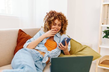 Rahat oturma odasında dizüstü bilgisayar ve kulaklık kullanan mutlu genç bir kadın, evden çalışıyor.