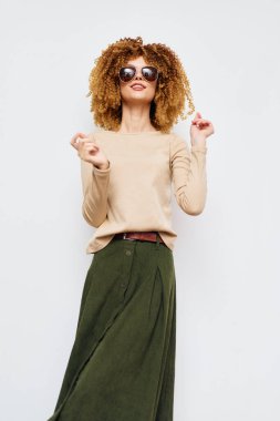 Kıvırcık saçlı, açık bej bluzlu ve akıcı yeşil etekli, temiz beyaz bir arka plana karşı poz veren, modern moda fotoğrafçılığı için mükemmel bir modaya uygun, eğlenceli ve modaya uygun bir hava yayan modaya uygun bir kadın model.