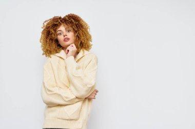 Hafif, büyük bir süveter giyen kıvırcık saçlı genç bir kadın, sade beyaz bir arkaplanda duruyor. Poz, moda ve yaşam tarzı için ideal bir güven ve oyunculuk karışımı sunuyor.