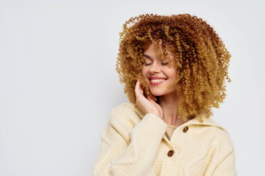 Yumuşak, krem renkli süveter giyen neşeli, kıvırcık saçlı genç bir kadın nazikçe yüzüne dokunuyor ve gülümsüyor, minimalist bir ışık ayarına neşe ve güven veriyor.