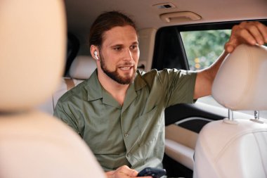 Uzun saçlı genç bir adam arabanın arkasında oturuyor, akıllı telefonuna odaklanmış, kablosuz kulaklık takıyor, açık renkli bir iç mekanı var.