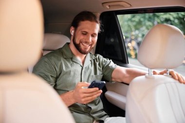 Neşeli bir adam arabanın arka koltuğunda eğleniyor, akıllı telefonuna odaklanmış, kablosuz kulaklık takıyor.