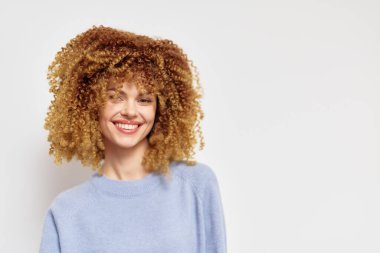 Yumuşak, kıvırcık saçlı, mutlu bir kadın yumuşak bir kazak giyiyor, pozitiflik yayıyor açık arka plan onun neşeli tavırlarını vurguluyor, moda ya da sağIık kavramlarına uygun hale getiriyor.