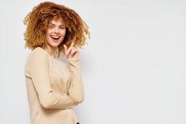 Kıvırcık saçlı neşeli bir kadın parlak bir arka planda gülümsüyor, neşeli bir hareketle elini kaldırıyor görüntü neşe ve güveni yakalıyor, pozitif bir yaşam tarzını yansıtıyor.