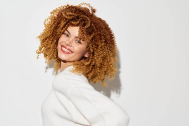 Şehvetli kıvırcık saçlı neşeli genç bir kadın, beyaz bir süveter içinde gülümsüyor, temiz beyaz bir arkaplanda duruyor, bireyselliği, mutluluğu ve kendini ifade edişini vurguluyor ve kutluyor.