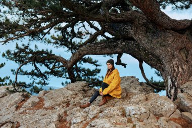 Sarı paltolu bir kadın kırsaldaki sakin bir ağacın gölgesinde, büyük bir kayanın üzerinde huzur içinde oturuyor.