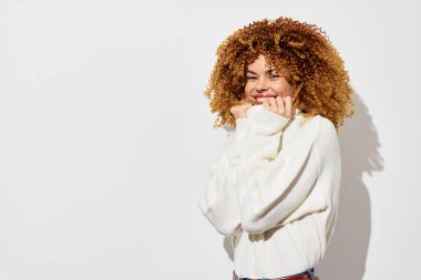 Kıvırcık saçlı neşeli bir kadın, sade beyaz bir arka plana sıcak bir şekilde gülümser, rahat bir süveter giyer, mutluluk saçar, pozitifliği ve doğal güzelliği somutlaştırır.