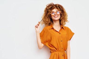 Kıvırcık saçlı ve turuncu elbiseli neşeli genç kadın beyaz bir arka plana karşı duruyor, kendine güveni ve mutluluğu yayıyor, moda, yaşam tarzı ve fotoğrafçılıkta olumlu duygular için ideal.
