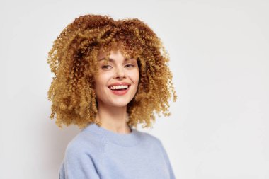 Şehvetli kıvırcık sarı saçları olan, açık mavi süveter içinde geniş bir gülümsemeye sahip neşeli genç bir kadın basit beyaz bir arkaplanda duruyor, pozitif ve sıcak, neşeli ve hayat dolu olmak için mükemmel.