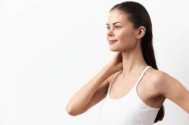 Pürüzsüz tenli, uzun saçlı, beyaz kolsuz tişörtlü, çarpıcı bir genç kadın zarif bir şekilde beyaz bir arka plana karşı duruyor, kendine güven ve sağlık yayıyor, güzellik ve refah teması için ideal.