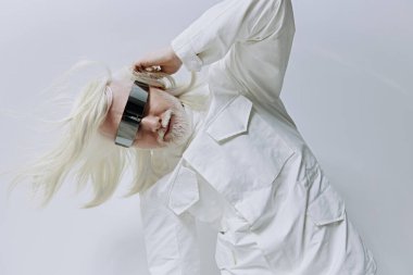 Uzun beyaz saçlı şık bir birey parlak siyah bir güneşlik takarken dramatik bir şekilde poz veriyor. Tarafsız gri arka plan çarpıcı bir kontrastı artırıyor, modernliği ve yaratıcılığı vurguluyor.