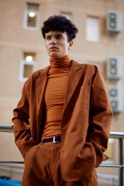 Kahverengi takım elbise ve balıkçı yaka giyen şık bir genç adam bulanık bir şehir geçmişine karşı poz veriyor.