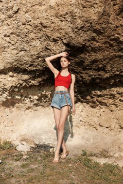 Kırmızı bluzlu ve kot şortlu kadın seyahat macerası sırasında heybetli kaya oluşumunun önünde duruyor.