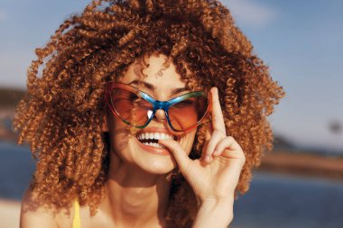 Şehvetli kıvırcık saçlı neşeli bir kadın güneş gözlüklerini açık havada tutarken gülümsüyor, parlak ve kaygısız bir yaz havası sergiliyor.