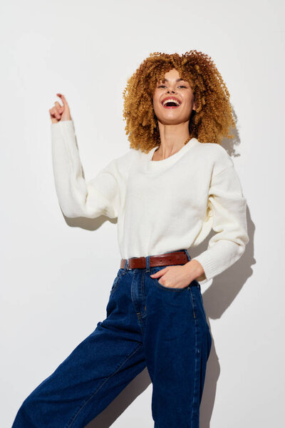 Веселая молодая женщина с объемными вьющимися волосами стоит на обычном белом фоне Она одета в простой белый свитер и высокие талии синие джинсы, улыбаясь и указывая вверх Thi