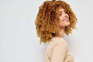 Bu görüntüde güzel kıvırcık saçlı bir kadın var. Yan tarafa bakarken hafifçe gülümsüyor. Yumuşak tarafsız arkaplan onun özelliklerini geliştiriyor.