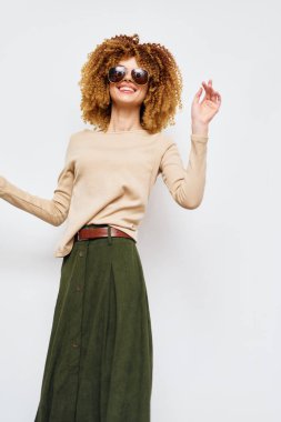 Kıvırcık saçlı, şık bej kazaklı ve yeşil etekli, şık güneş gözlüğü takan, minimalist beyaz bir fona karşı neşeli bir poz veren mutlu bir kadın.