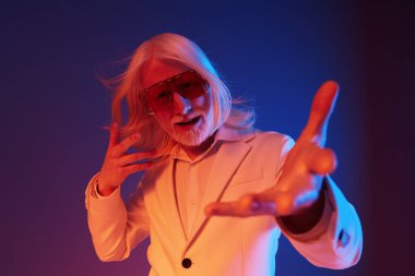 Uzun beyaz saçlı, güneş gözlüğü takan ve beyaz takım elbiseli şık, yaşlı bir adam, ifadesindeki kendine güven ve karizmayı gözler önüne seren canlı mavi ve pembe bir arkaplan karşısında dramatik bir şekilde poz veriyor.