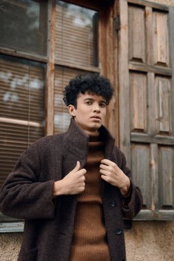 Kahverengi ceketli ve kaplumbağa yakalı şık bir genç arka planda penceresi olan eski bir ahşap kapının önünde duruyor..