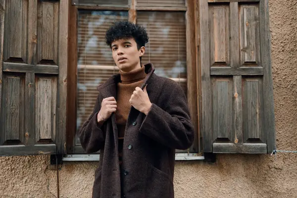 Kahverengi ceketli ve balıkçı yaka kazaklı şık bir genç, pencerenin önünde duruyor..