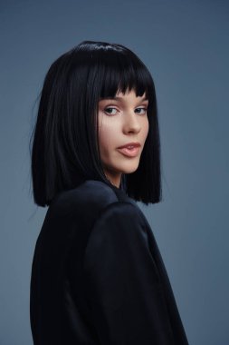 Siyah ceketli, kısa siyah saçlı güzel bir kadın, mavi arka planda duruyor, kameraya özgüvenle bakıyor, modern moda ve kişisel tarzını sergiliyor.