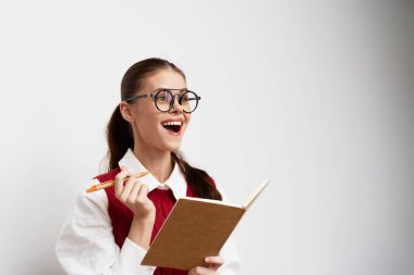 Geniş gözlüklü neşeli genç bir kadın elinde bir defter ve kalem, geniş bir gülümsemeyle minimalist arkaplan, öğrenme ve yazmada ilham ve yaratıcılığı sembolize ederek onun ifadesini vurguluyor.