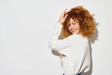 Kıvırcık saçlı neşeli bir kadın parlak bir arka planda neşeli bir poz sergiliyor. Parlak gülüşü ve rahat süveteri davetkar ve neşeli bir atmosfer yaratıyor. Güzelliği ve güveni ön plana çıkarıyor.