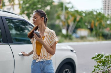 Genç bir kadın, canlı bir şehir ortamında, modern beyaz bir aracın önünde duran bir telefon görüşmesinde, arka planda yemyeşil ve parlak şehir yapıları yer alıyor.