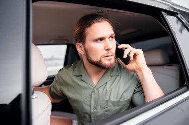 Sakallı ve uzun saçlı bir adam arabada oturuyor, telefonla konuşuyor, konsantrasyonunu gösteriyor.