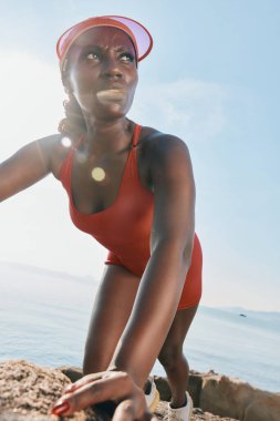 Kırmızı mayo ve şapkalı güçlü, atletik bir kadın, su kenarında kayalık bir alana tırmanır. Güneş ışıl ışıl parlar ve arka planda açık mavi bir gökyüzü vardır.