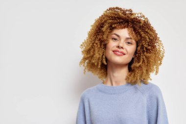 Şehvetli kıvırcık sarı saçlı neşeli genç bir kadın yumuşak mavi bir süveter giyerken sıcacık gülümserken doğal güzelliğini ve pozitifliğini vurgulayan basit bir beyaz arkaplan içerir.