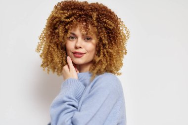 Bu resim, yumuşak mavi bir süveter giymiş, şehvetli, kıvırcık saçlı genç bir kadını gösteriyor. Hafif, nötr bir arkaplanda poz veriyor, rahat ama kendine güvenen bir ifade yayıyor.