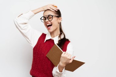 Gözlüklü neşeli genç bir kadın bir elinde bir defter, diğer elinde alnına dokunuyor. Beyaz bir gömleğin üzerine kırmızı bir kazak giyiyor. Tarafsız bir arkaplana yaslanmış.