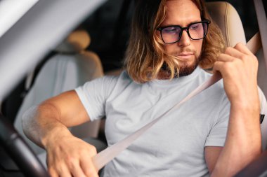 Uzun saçlı ve gözlüklü genç bir adam modern bir arabada otururken emniyet duygusunu gösteriyor.