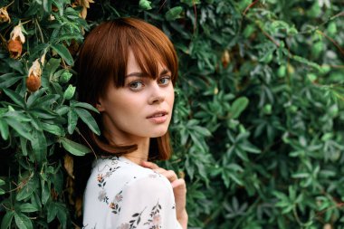 Kızıl saçlı, dingin bir kadın yemyeşil bitkilerin yanında dikiliyor, uzaklara bakıyor.