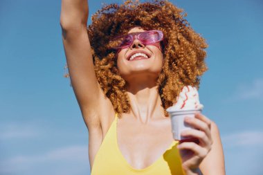 Kıvırcık saçlı ve parlak güneş gözlüklü neşeli genç bir kadın, arka planda parlak mavi gökyüzü ile güneşli havada kutlama yaparken elinde bir dondurma külahı tutuyor.
