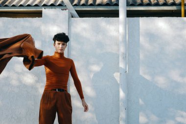 Kahverengi balıkçı yaka ve pantolon giyen modaya uygun bir erkek model, parlak beyaz bir duvara dayalı, dinamik bir kumaş parçasıyla poz veriyor.