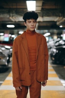 Pas rengi ceket ve balıkçı yaka kazak giymiş genç bir adam loş bir otoparkta güvenle poz veriyor.