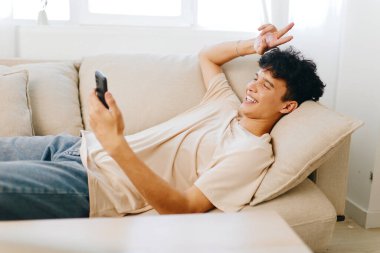 Neşeli genç bir adam açık renkli bir kanepede uzanıyor, akıllı telefonuyla meşgul olurken bir barış işaretiyle şakalaşıyor parlak ve havadar oturma odası arka planı rahatlamış havayı arttırıyor.