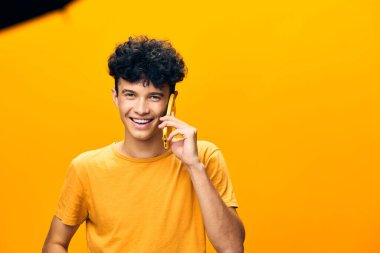 Elinde telefon tutan, gülen ve muhabbete dalan, günlük turuncu bir tişört giyen genç adam parlak sarı arka plan neşeli tavrını ve pozitif enerjisini artırıyor.