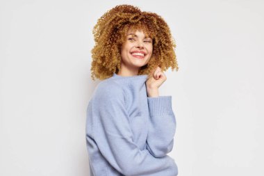 Şehvetli kıvırcık saçlı, yumuşak, açık mavi bir kazak giyen neşeli bir genç kadın. Sıcak bir şekilde gülümsüyor. Sade, tarafsız bir geçmişe karşı güven ve mutluluk yayıyor.