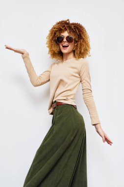 Sarışın, kıvırcık saçlı, güneş gözlüklü, bej renkli, uzun kollu, yeşil etekli, minimalist, hafif bir arka plana sahip, kendine güvenen ve oynayan bir kadın manken.