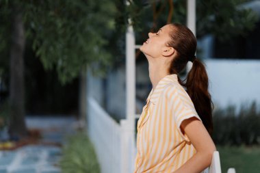 Açık havada duran neşeli genç bir kadın, güneşlenirken, hoş bir ifadeyle açık çizgili bir gömlek giyiyor, yemyeşil ve rahatlatıcı bir atmosferle çevrili.