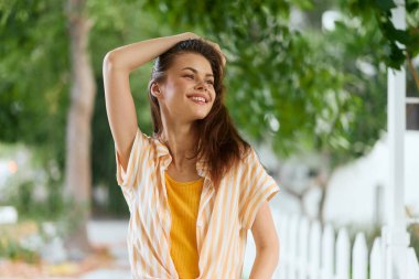 Uzun saçlı neşeli genç kadın, rahat bir şekilde dışarıda poz veriyor. Görüntü arka planda canlı yeşil yapraklar içeriyor, taze ve canlı bir atmosfer ekliyor.