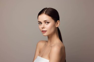 Beyaz askısız elbiseli genç bir kadın gri bir arka plana karşı kameraya poz veriyor..