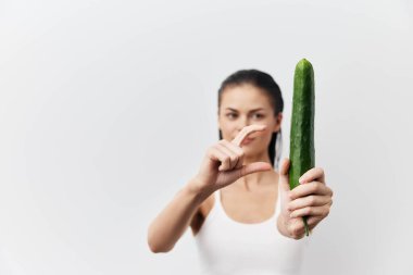 Beyaz arka planda salatalık tutan oyuncu bir kadın, mizahi boyutta bir karşılaştırmayı vurguluyor tazelik ve canlılık yayıyor, sağlıklı yaşam tarzı temaları için mükemmel.