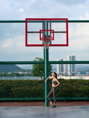 Atletik kadın basketbol potasının yanında duruyor, modern şehir arka planına karşı fitness yaşam tarzını sergiliyor