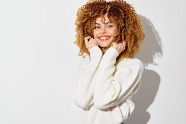 Kıvırcık saçlarını gösteren, rahat beyaz bir kazak giyen gülümseyen genç bir kadın. Parlak, temiz bir arkaplana karşı şakacı bir şekilde poz veriyor. Olumlu ve özgüven yayıyor.