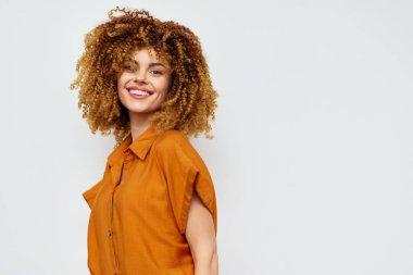 Şehvetli kıvırcık saçlı, neşeli bir kadın. Topraksı kahverengi bir gömlek giyiyor. Temiz, açık bir arkaplana yaslanmış.